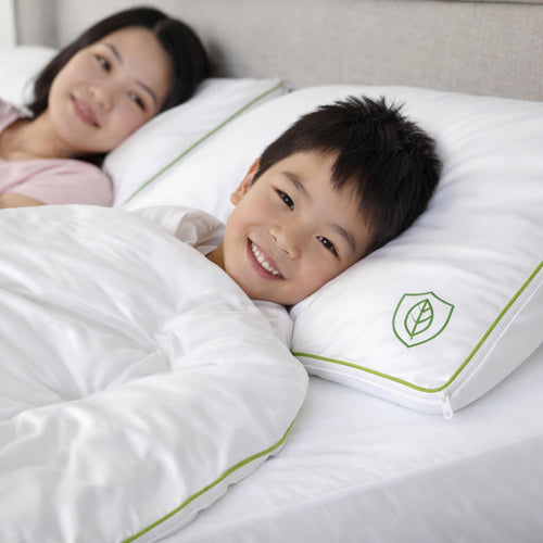 Child and woman lying on a bed with a white comforter and pillow featuring a green leaf logo with no dust mite allergy